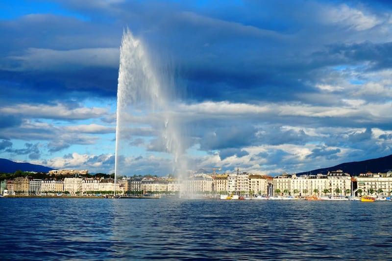 A view on the city of Geneva in the sunshine with clouds at dusk and the Jet d'Eau, located in the middle of Lake Geneva at the city of Geneva, Switzerland. The current Jet d'Eau was inaugurated in 1951. Reflections in the water.