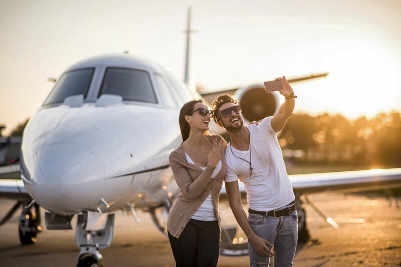 Young urban heterosexual couple with sunglasses standing at the airport track in front of private jet airplane and posing for a selfie. Man is holding the camera and taking a picture. Bright sunlight is in the background.