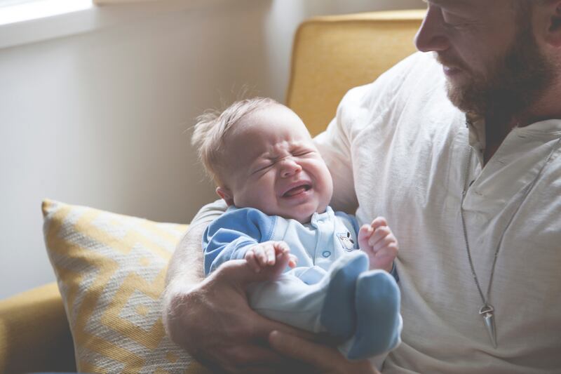 Father holding and looking at crying baby, sitting on sofa. Getty Images