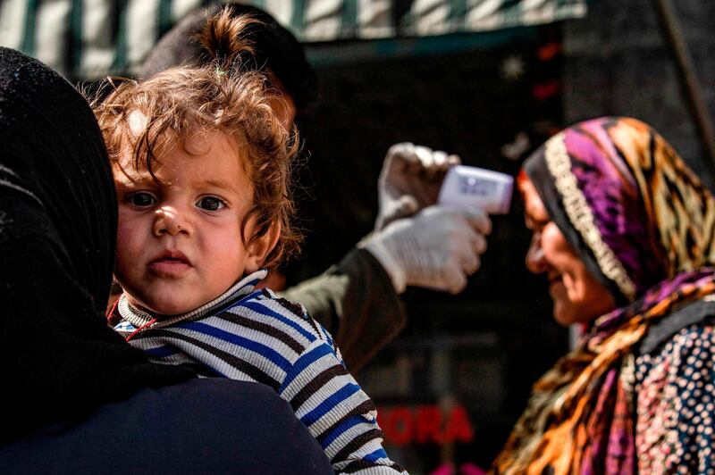 A child looks on as a member of the Kurdish Internal Security Forces of Asayesh checks a woman's temperature in the Kurdish-majority city of Qamishli in Syria's northeastern Hasakah province, on March 23, 2020, amid measures to curb the spread of the novel coronavirus. The Kurdish authorities in northeast Syria have not recorded any deaths so far, but have imposed a curfew in a bid to stem any outbreak. / AFP / DELIL SOULEIMAN
