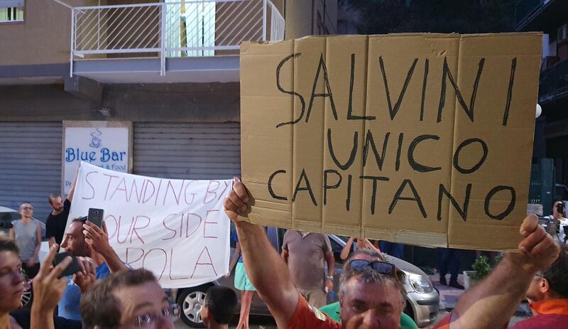 A man holds a placard reading "Salvini only captain" (R) as others hold a banner reading "Standing by your side Carola" (Rear L) as people and the media gather outside the building where Sea-Watch 3 captain Carola Rackete has been staying under house arrest, on July 2, 2019 in Agrigento, Sicily, after an Italian judge said she was free to go, three days after her arrest for docking with 40 migrants aboard her rescue ship in defiance of an Italian ban. German Rackete was arrested after hitting a police speed boat while entering the port of the southern island of Lampedusa in her vessel, banned from docking by Italian authorities. / AFP / Giovanni ISOLINO
