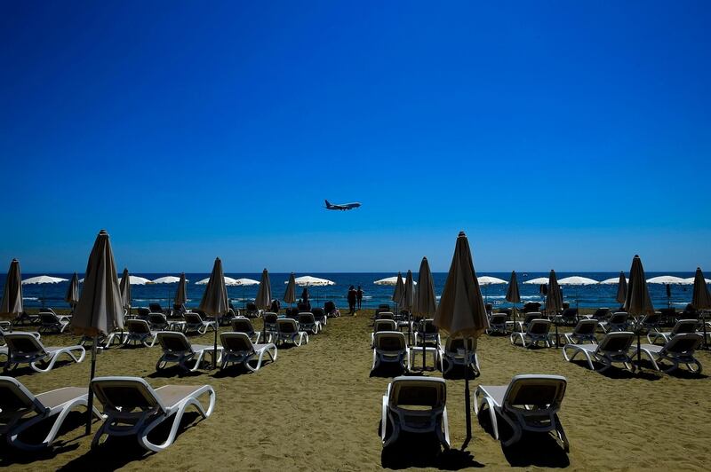 People stand by the sun-beds and umbrellas as an aircraft prepares for landing at Larnaca international airport, at Makenzi beach in southern city of Larnaca, Cyprus, Tuesday, May 18, 2021.  Tourism accounts directly for 13 percent of Cyprus' gross domestic product and authorities are striving to attract mass arrivals by touting the country's decision for quarantine requirements to holiday makers from 65 countries. (AP Photo/Petros Karadjias)