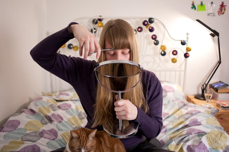 A young blonde woman cuts her hair at home using a hand mirror. She sits on her bed with her pet cat sitting on her lap.