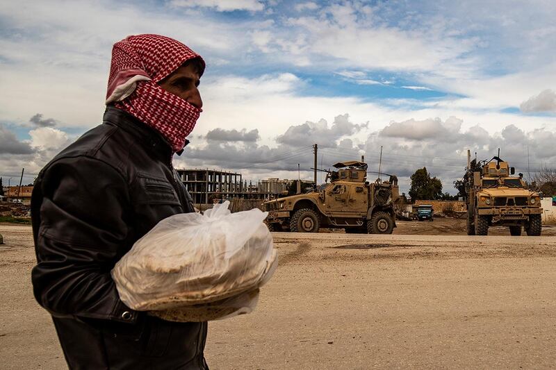 A man carrying bags of bread walks as a patrol of US military vehicles is seen in the town of Tal Tamr in the northeastern Syrian Hasakeh province along the border with Turkey on February 8, 2020. (Photo by Delil SOULEIMAN / AFP)