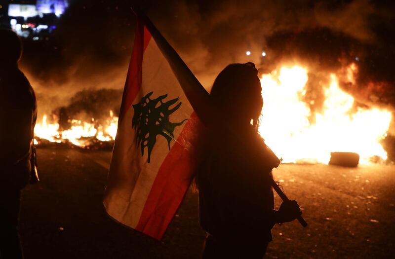 A Lebanese anti-government protester carries a Lebanese flag in front of burning tires blocking the main highway linking the city of Tripoli to Beirut at the coastal city of Byblos, north of the capital. AFP