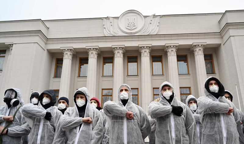 Activists of National Corps far-right party wearing gas masks and protective suits  salute as they together with different Ukrainian opposition parties supporters rally in front of the parliament in Kiev during its extraordinary session on March 17, 2020, demanding to stop the parliament's work for quarantine and not to consider the laws ambiguously perceived in society, while limits on mass gatherings over coronavirus fears are on place. / AFP / Sergei SUPINSKY

