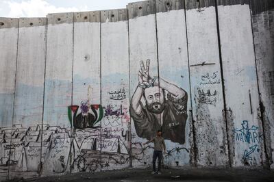 A Palestinian man tries to hitch a ride as he stands below the giant graffiti showing imprisoned political figure Marwan Barghouti , Palestinian President Abbas and UN tents painted on Israel's  8 meter high cement separation barrier on the West Bank side of the divided Palestinian village of Abu Dis .(Photo by Heidi Levine for The National).
