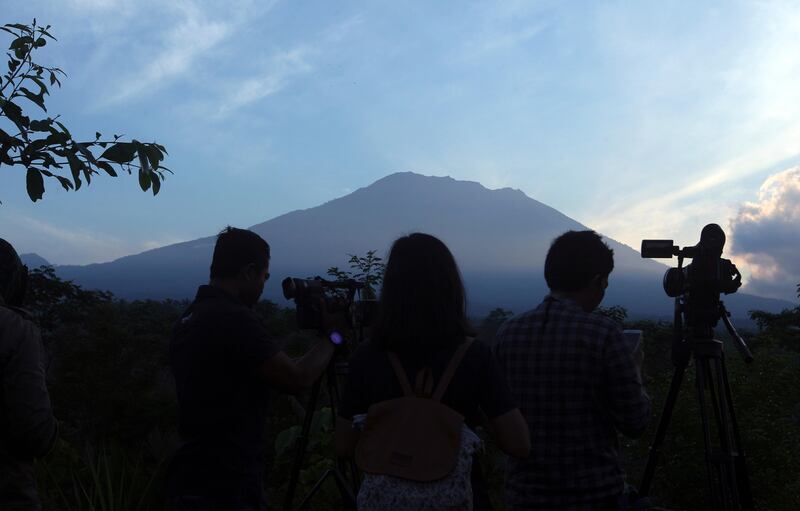 Journalists watch Mount Agung from an observation point which is about 12 kilometer (7.4 miles) away from the volcano in Karangasem, Bali, Indonesia, Thursday, Sept. 28, 2017. The exodus from the menacing volcano on the Indonesian tourist island is nearing 100,000 people, a disaster official said Wednesday, as hundreds of tremors from the mountain are recorded daily. (AP Photo/Firdia Lisnawati)