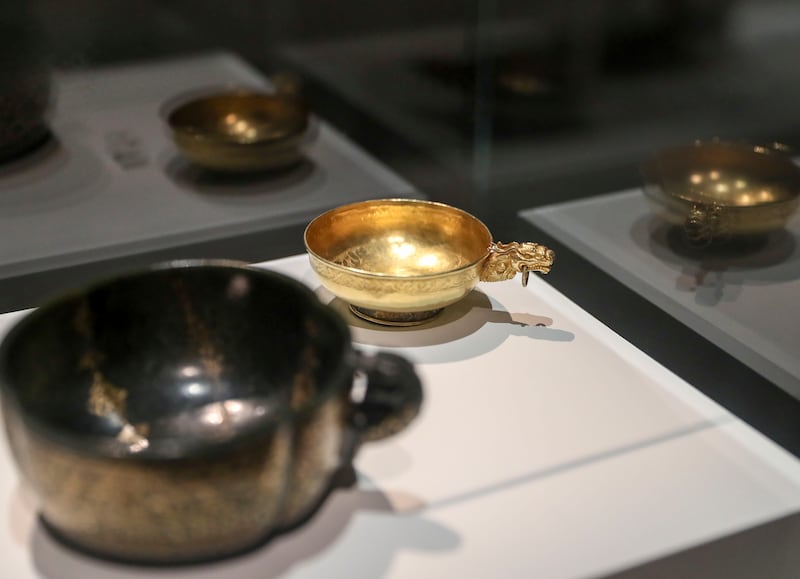 Right, a cup with dragon-shaped handle from China, Yuan dynasty (1279-1368) in hammered embossed gold with chasing. Left, a larger, more bulbous variation made of black jade.  Khushnum Bhandari / The National