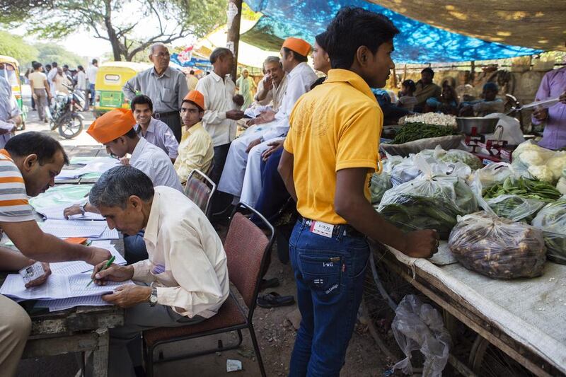 A customer waits at a vegetable stall while polling officials check the details of voters at a polling station during the third phase of election voting in New Delhi, India, on Thursday, April 10, 2014. Prashanth Vishwanathan/Bloomberg