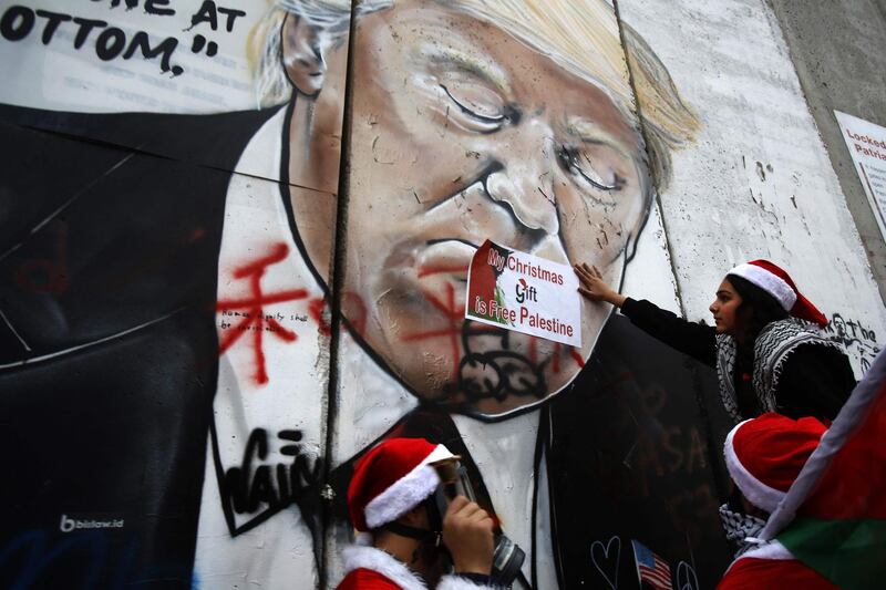 TOPSHOT - Palestinians dressed as Santa Claus, hang signs on a mural painting of US President Donald Trump on Israel's controversial separation barrier in the West Bank city of Bethlehem, on December 23, 2018. / AFP / Musa AL SHAER
