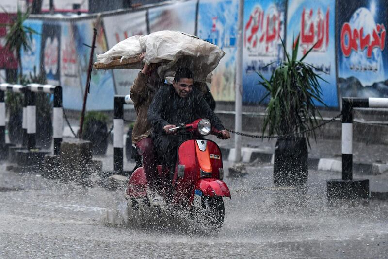 A man carries a wooden board loaded with loaves of bread, covered with plastic, while riding in the back of a scooter amidst a heavy rain storm. AFP