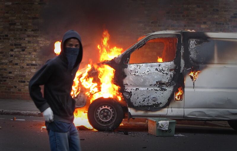 A hooded rioter walks past a burning vehicle in Hackney.