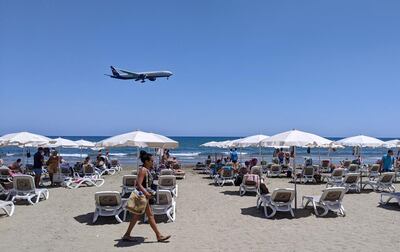 A passenger plane approaches Larnaca airport before landing as a woman walks past sunbeds at Makenzy Beach in the southern Cypriot port city on May 23, 2021. A 39-year-old British woman died in a Cypriot hospital as a result of  a blood clotting incident after receiving the AstraZeneca Covid-19 vaccine, the official Cyprus News Agency said May 24, 2021. / AFP / Etienne TORBEY
