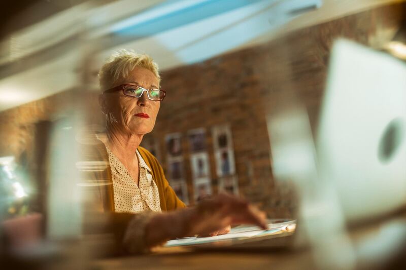 Mature adult businesswoman writing on paper and using a laptop at night.