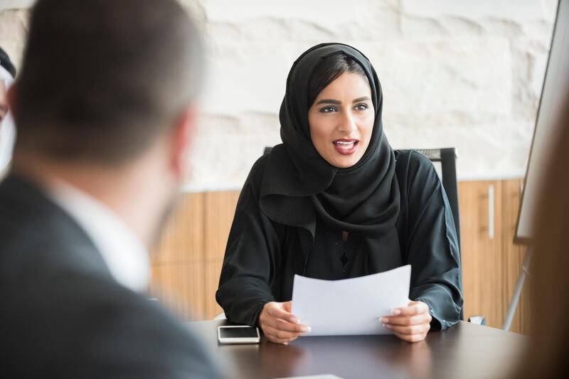 A photo of mature businessman looking at colleague. He is wearing traditional clothes. Professional is sitting and smiling at coworkers. Office workers are in a meeting, at brightly lit workplace.