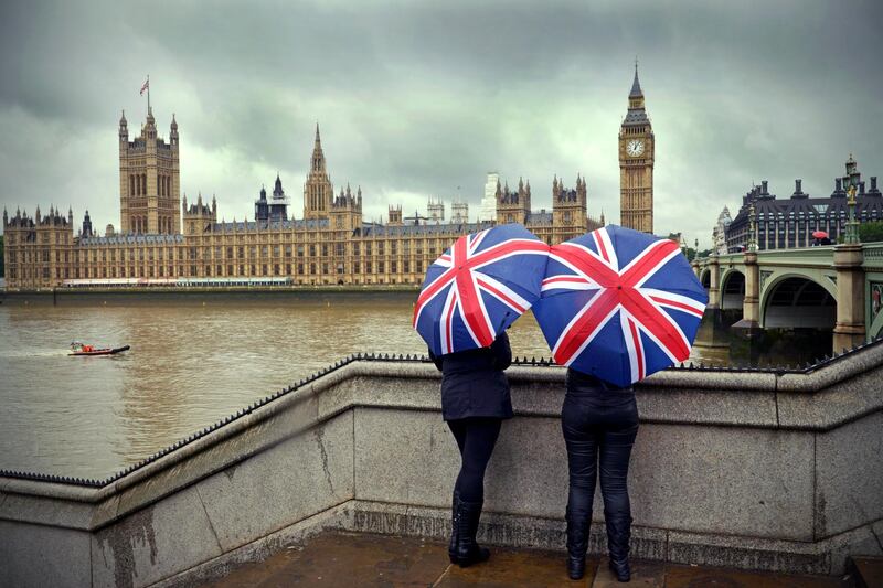 "Tourists huddle beneath British flag umbrellas (they sell them there) during a London summer rainstorm near the Houses of Parliament. A dark cloudy typical summer day!Westminster, LondonUK"