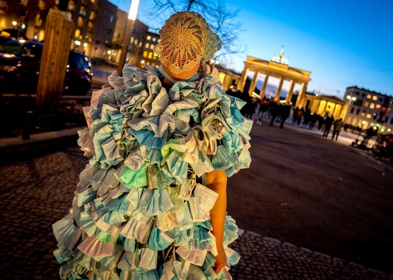 A woman wears a dress made of hundreds of face masks as she returns from an anti- coronavirus demonstration in Berlin.  In the background is the Brandenburg Gate.  AP