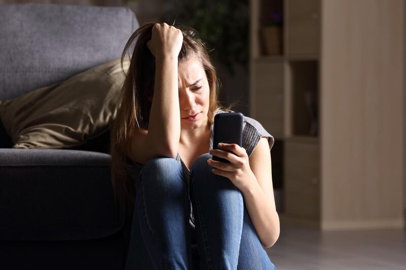 JB029B Front view of a sad teen checking phone sitting on the floor in the living room at home with a dark background