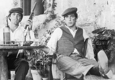 1972:  American actor Al Pacino (right) sits with Italian actor Franco Citti holding a rifle at an outdoor table in director Francis Ford Coppola's film, 'The Godfather'. Pacino rests his leg on a chair.  (Photo by Paramount Pictures/Getty Images)