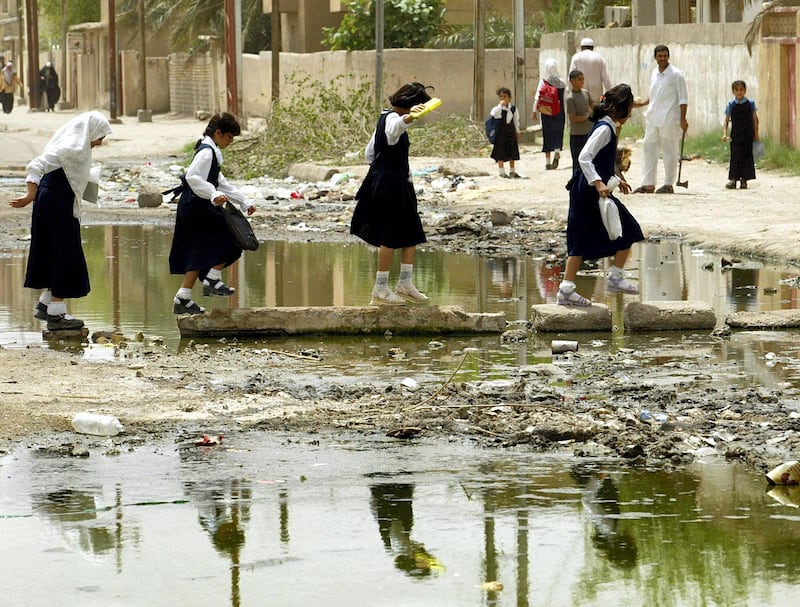 Young school girls cross a trash ridden patch of water on stones in the middle of the road on their way home in the southrn city of Basra 18 May 2003. The pool of dirty water has been on the street off and on for the last ten years and according to local residence has caused them to become ill. AFP PHOTO/Timothy A. CLARY (Photo by TIMOTHY A. CLARY / AFP)