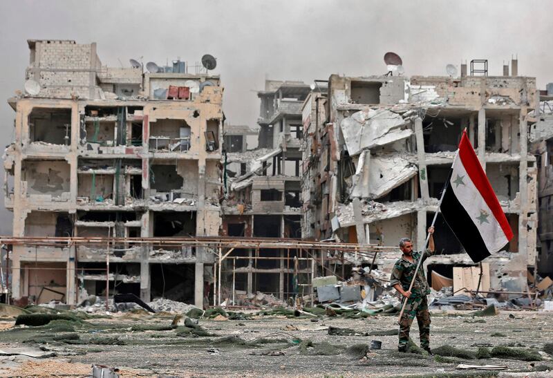 CORRECTION / TOPSHOT - A member of the Syrian pro-government forces carries the national flag as he stands in front of damaged buildings in the Yarmuk Palestinian refugee campon the southern outskirts of the capital Damascus on May 22, 2018.  The government seized earlier in the week the Yarmuk Palestinian camp and adjacent neighbourhoods of Tadamun and Hajar al-Aswad from the Islamic State (IS) group , putting Damascus fully under its control for the first time since 2012. / AFP / LOUAI BESHARA
