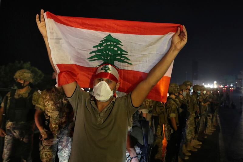 An anti-government protester holds up a Lebanese flag as army soldiers stand guard during a demonstration against deteriorating economic conditions as politicians are deadlocked over forming a new government, in the town of Jal el-Dib, north of Beirut, Lebanon, Sunday, Sept. 27, 2020. Lebanese Prime Minister-designate Mustapha Adib resigned Saturday amid a political impasse over government formation, dealing a blow to French President Emmanuel Macron's efforts to break a dangerous stalemate in the crisis-hit country. (AP Photo/Bilal Hussein)