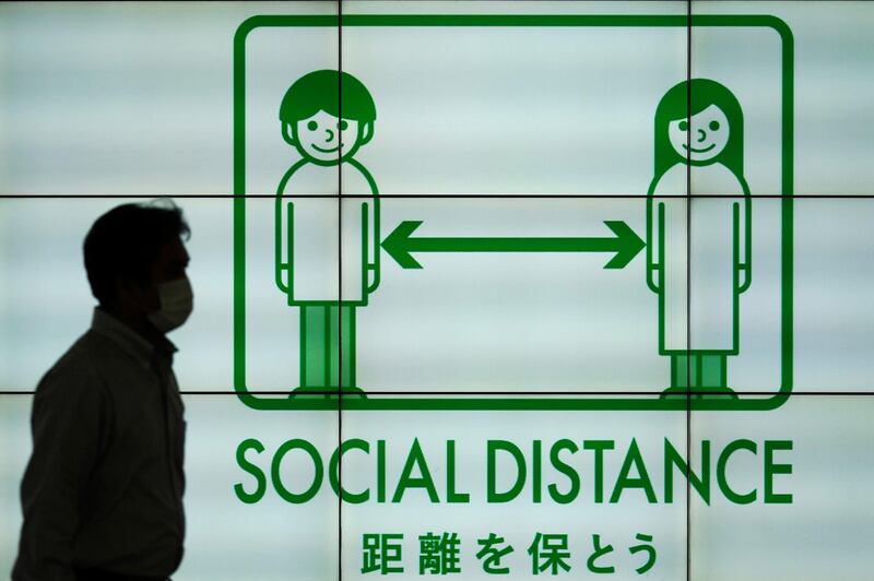 A man wearing a face mask to help curb the spread of the coronavirus walks in front of a banner reminding the public of social-distancing rules in Tokyo. AP Photo