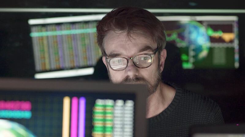 A bearded man studying computer displays depicting global trends with figures, the screens being reflected in his glasses as well as behind him on a black glass wall. The subject matter on the screens pertains to ecology, economics and global trends.