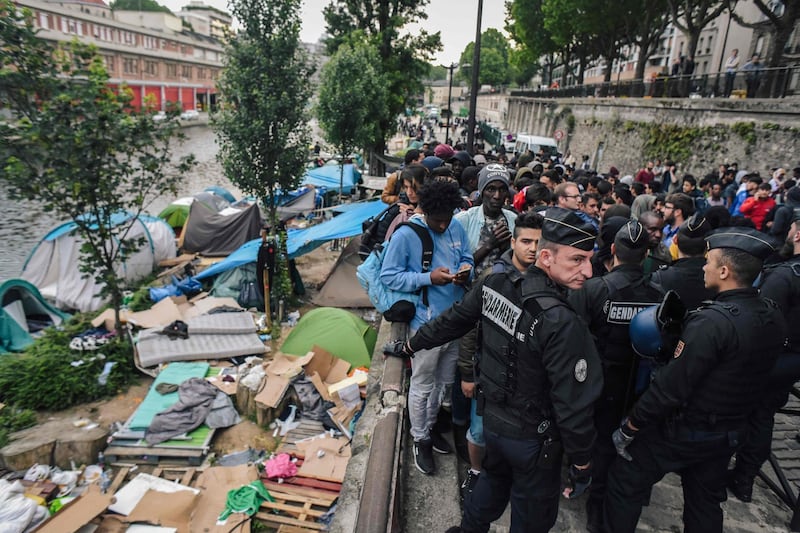 TOPSHOT - French CRS anti-riot police officers stand guard by migrants and refugees queing during the evacuation by police of their makeshift camp along the Canal de Saint-Martin at Quai de Valmy in Paris, on June 4, 2018. More than 500 migrants and refugees were evacuated on early June 4, 2018 from a makeshift camp that had been set up for several weeks along the Canal. / AFP / LUCAS BARIOULET
