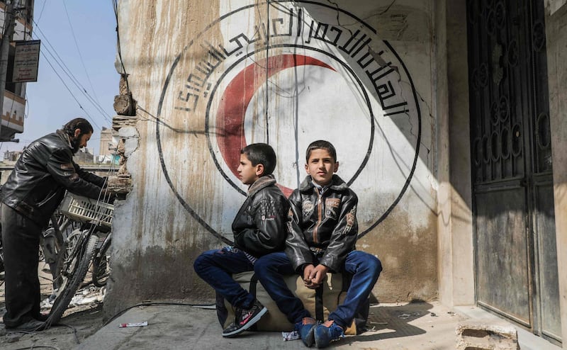 Syrian children sit before a mural on a wall bearing the logo of the Syrian Red Crescent in the Syrian town of Douma in the rebel-held enclave of Eastern Ghouta on the eastern outskirts of the capital Damascus on March 13, 2018. AFP