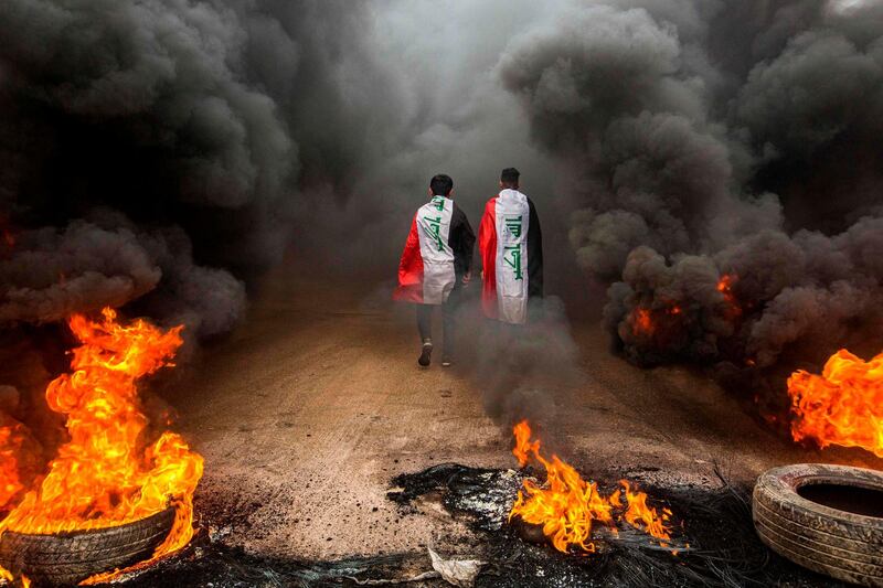 TOPSHOT - Anti-government protesters draped in Iraqi national flags walk into clouds of smoke from burning tires during a demonstration in the southern city of Basra on November 17, 2019, as protesters cut-off roads and activists call for a general strike. Iraqis flooded the streets of the capital and southern cities in a general strike that bolstered the weeks-long movement demanding a government overhaul. Protesters cut roads in the oil-rich port city of Basra by burning tyres and in Hillah, south of Baghdad, students and other activists massed in front of the provincial headquarters. / AFP / Hussein FALEH
