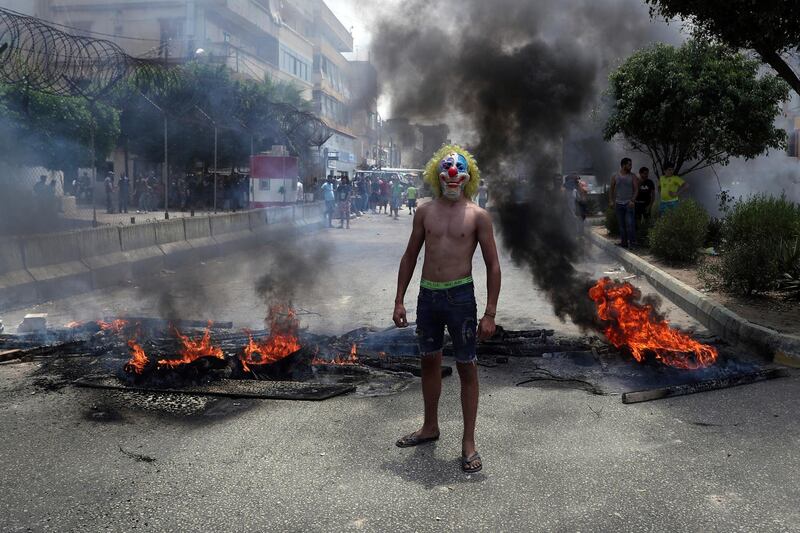 An anti-government demonstrator poses in a clown mask as others burn tires and wood to block a road in Beirut, Lebanon. AP