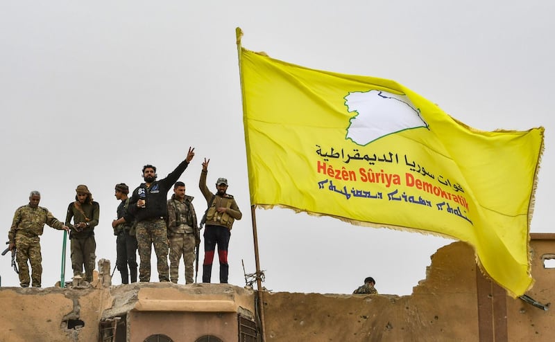 Fighters of the Syrian Democratic Forces (SDF) flash the victory gesture next to their unfurled flag atop a roof at a position in the village of Baghouz in Syria's eastern Deir Ezzor province near the Iraqi border on March 24, 2019, a day after the Islamic State (IS) group's "caliphate" was declared defeated by the US-backed Kurdish-led SDF.  / AFP / GIUSEPPE CACACE
