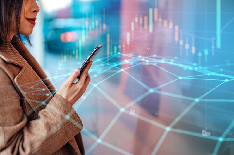 Closeup shot of young woman analysing and checking stock market over smartphone in downtown financial district. Stock exchange market trading board in background. Blockchain concept. Crytocurrency market.