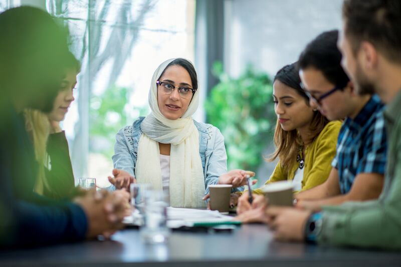 A multi-ethnic group of business people are indoors in an office boardroom. They are sitting around a table and making plans. A woman wearing a head scarf is explaining papers to her coworkers.