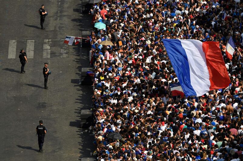 Security forces stand guard by supporters waving French national flags and gathering at the Place Charles de Gaulle up the Champs-Elysees avenue in Paris on July 16, 2018 as they wait for the arrival of the French national football team for celebrations after France won the Russia 2018 World Cup final football match on the previous night. / AFP / Bertrand GUAY
