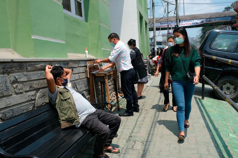 A man rests on a bench as pedestrian wash their hands at hand basins set up by the entrance of Oking Hospital in Kohima, capital of the northeastern Indian state of Nagaland, Monday, April 13, 2020. A man from Nagaland state has tested positive for COVID- 19, becoming the first case from the state. The new coronavirus causes mild or moderate symptoms for most people, but for some, especially older adults and people with existing health problems, it can cause more severe illness or death. (AP Photo/Yirmiyan Arthur)
