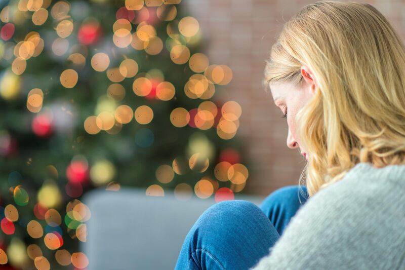 A Caucasian woman is indoors in her living room. There is a Christmas tree in the background. The woman is wearing warm clothing. She is sitting on the couch and looking sad because she is alone on Christmas day.