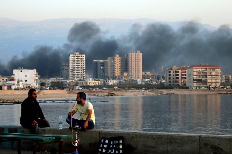 Lebanese men smoke shisha as smoke billows over the northern Lebanese city of Tripoli on November 13, 2019, nearly a month into an unprecedented anti-graft street movement. Street protests erupted, the night before, after President Michel Aoun defended the role of his allies, the Shiite movement Hezbollah, in Lebanon's government. Protesters responded by cutting off several major roads in and around Beirut, the northern city of Tripoli and the eastern region of Bekaa. / AFP / Ibrahim CHALHOUB
