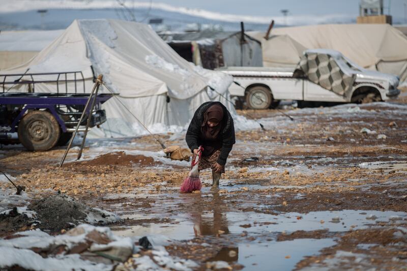 An elderly displaced Syrian woman removes snow in front of her tent.