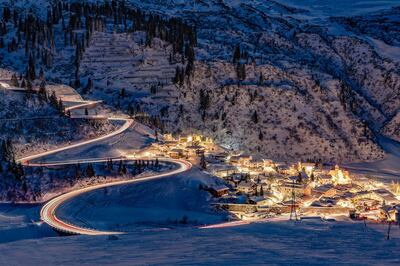 a winter scenery showing the village of Stuben am Arlberg in Vorarlberg at twilight. Light trails of cars driving up and down the windy mountain road are visible. The ground is covered with snow.