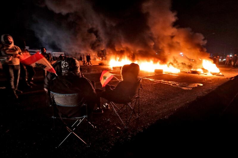 Anti-government protesters sit on chairs waving Lebanese national flags as tyres are aflame to cut off a section of the Beirut-Byblos highway in the coastal city of Byblos, as they protest the newly-announced government formed by Prime Minister Hassan Diab.  AFP
