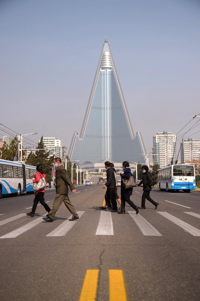 People wearing face masks walk across a street before the Ryugyong hotel (back C) on the occasion of the 108th birthday of late North Korean leader Kim Il Sung, known as the 'Day of the Sun', in Pyongyang on April 15, 2020. (Photo by KIM Won Jin / AFP)