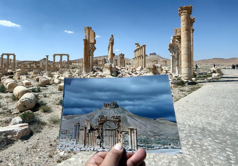 A general view taken on March 31, 2016 shows a photographer holding his picture of the Arc du Triomphe (Triumph's Arch) taken on March 14, 2014 in front of the remains of the historic monument after it was destroyed by Islamic State (IS) group jihadists in October 2015 in the ancient Syrian city of Palmyra. - Syrian troops backed by Russian forces recaptured Palmyra on March 27, 2016, after a fierce offensive to rescue the city from jihadists who view the UNESCO-listed site's magnificent ruins as idolatrous. (Photo by JOSEPH EID / AFP)
