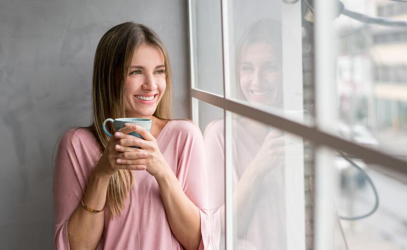 Casual woman at home drinking a cup of coffee and looking through the window. Getty Images