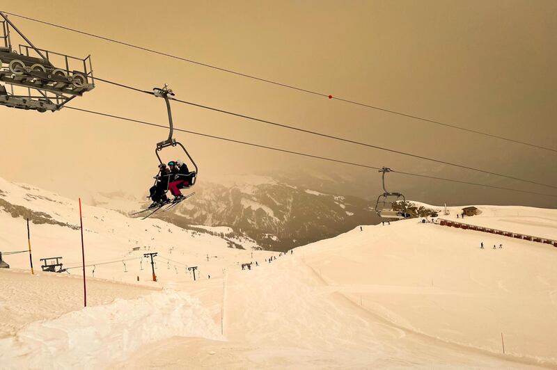 Skiers wearing protective face masks sit on a chairlift as Sahara sand colours the snow and the sky in orange and creates a special light atmosphere, during the coronavirus disease (COVID-19) outbreak, in the Alpine resort of Anzere, Switzerland, on Saturday, 6 February 2021. (Laurent Gillieron/Keystone via AP)