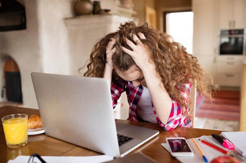 Upset teenage girl with long curly hair at home sitting at the table, working on notebook, studying. Getty Images