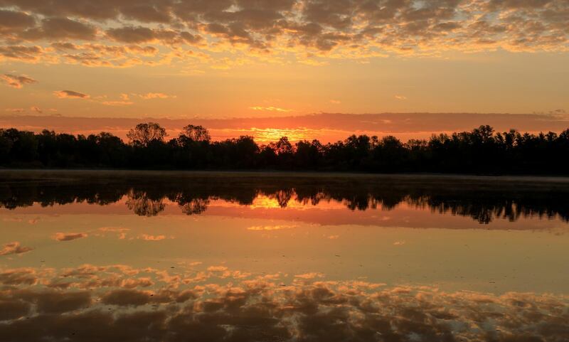 Clouds in the sky are reflected in the water below as the sun rises behind trees along the banks of a lake in Bruehl in Germany. EPA
