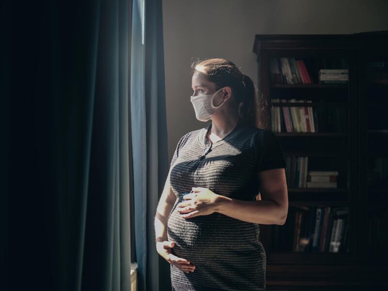 Pregnant woman with face mask standing in front of window.